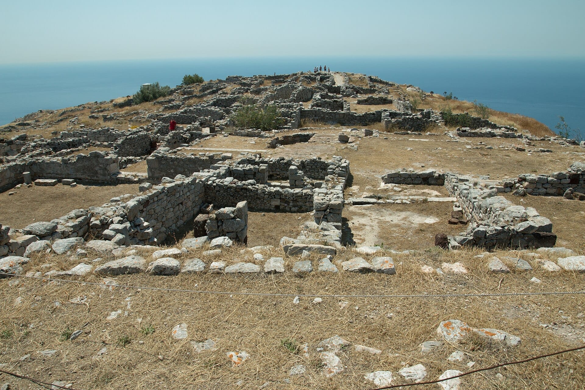 ซากปรักหักพังหินของ Ancient Thera ที่กระจายอยู่บนที่ราบสูงกลางแสงแดด มองเห็นทะเลอีเจียนสีน้ำเงินเข้มบนสันเขา Mesa Vouno ของ Santorini