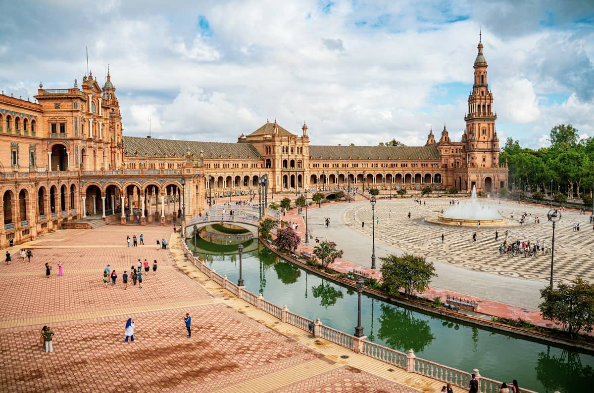 Plaza de España ที่เซบียา เห็นอาคารรูปครึ่งวงกลม หอคอยกลาง คลอง ต้นไม้ และผู้คนเดินข้ามจัตุรัสใต้ท้องฟ้ามีเมฆบางส่วน