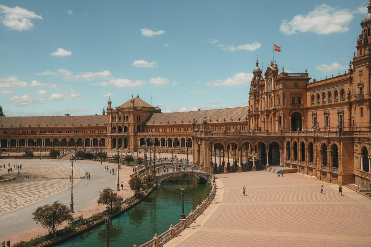 วิวกว้างของ Plaza de España ในเมือง Seville แสดงให้เห็นอาคารโค้ง หอคอย คลอง และผู้คนที่กำลังเดินอยู่ใต้ท้องฟ้าสดใส