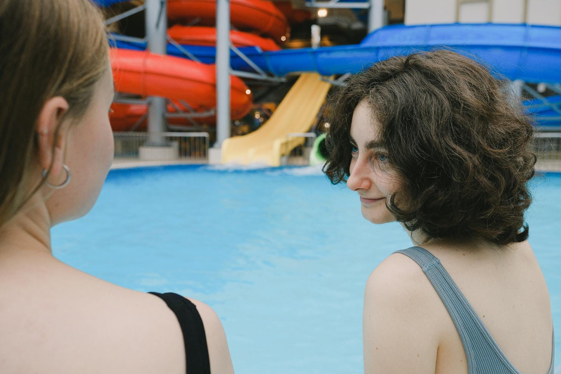Two women in swimsuits relax by a bright blue pool at Andamanda Phuket Water Park, with colorful slides visible in the background.