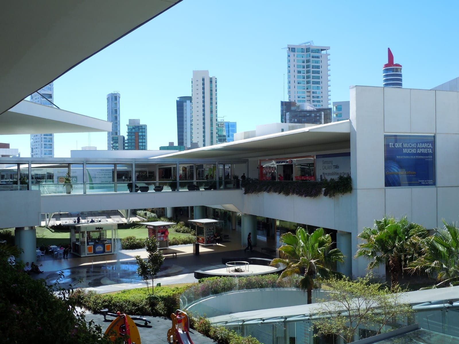 Architettura moderna e giardini rigogliosi all'Andares Shopping Mall a Zapopan, con grattacieli sullo sfondo di un luminoso cielo azzurro.