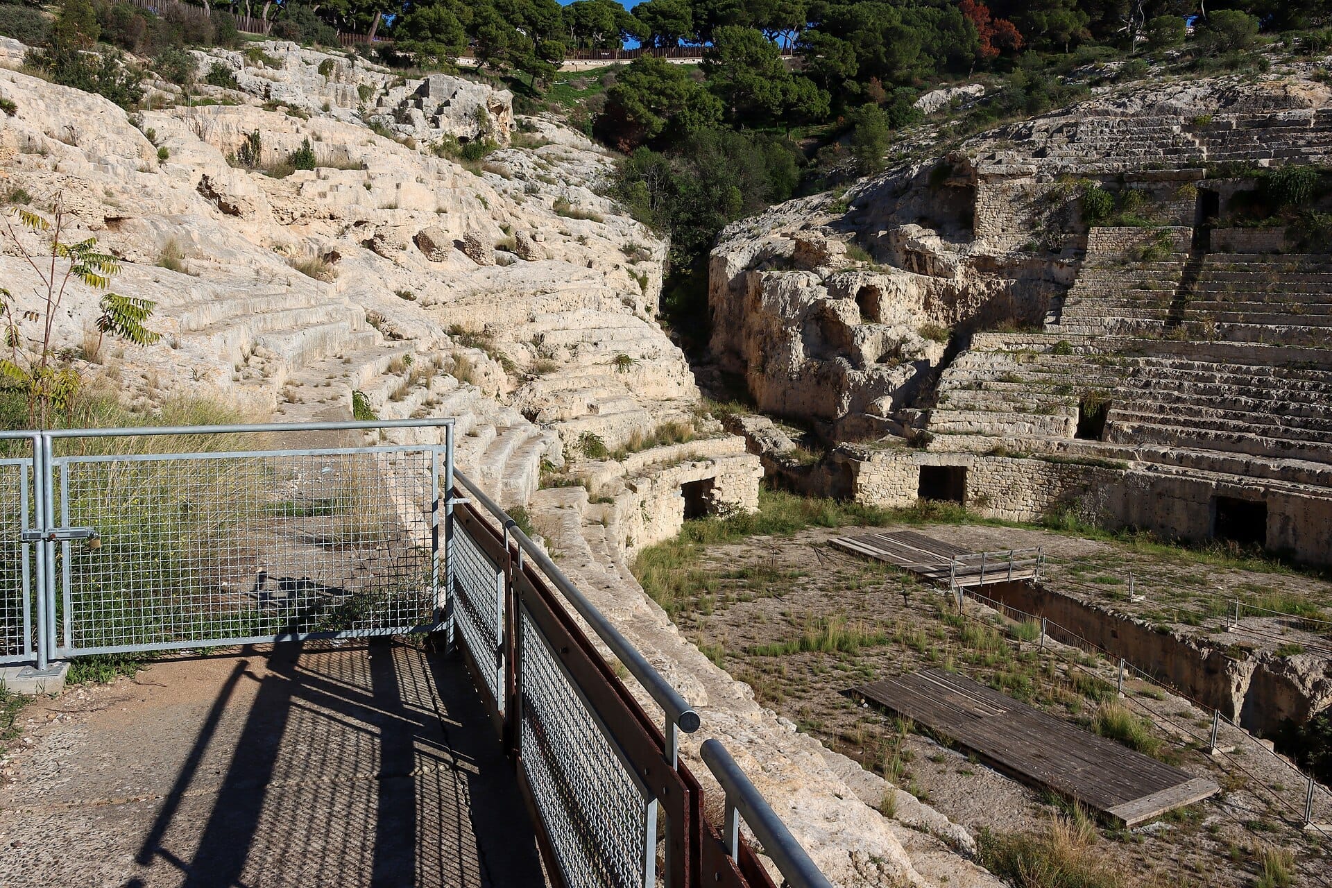 Blick auf das antike Römische Amphitheater von Cagliari mit verwitterten, in einen Hügel gemeißelten Steinsitzen und einem verschlossenen Metalltor im Vordergrund.