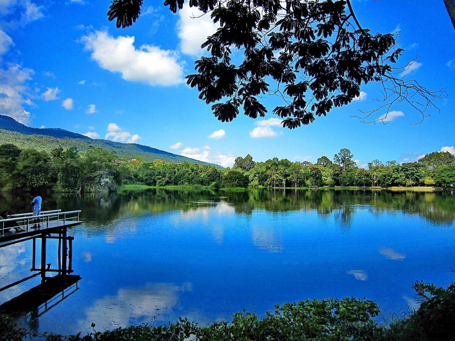 Hồ chứa nước Ang Kaew tại Đại học Chiang Mai được bao quanh bởi những cây xanh tươi tốt, hình ảnh phản chiếu rõ nét trên mặt hồ, núi non và bầu trời xanh ở phía sau.