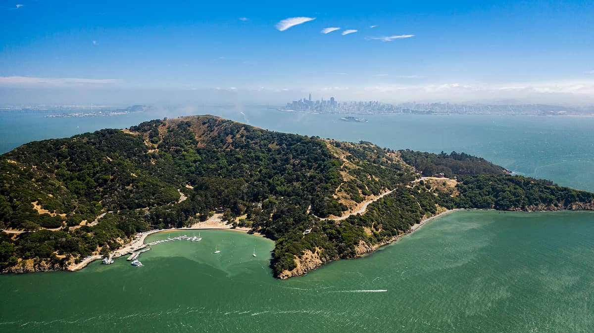 Vista aérea de Angel Island State Park rodeada por las aguas verde-azuladas de la bahía, con una marina y colinas boscosas, y el horizonte de San Francisco visible al fondo.