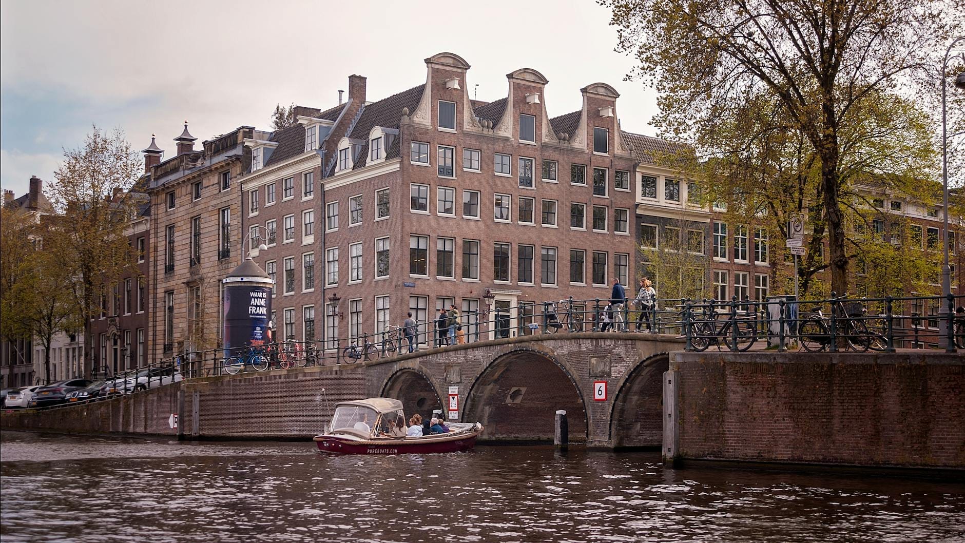 Vista amplia de la Casa de Ana Frank y el museo a lo largo del canal Prinsengracht en Ámsterdam, con un barco pasando bajo un puente histórico de arco.