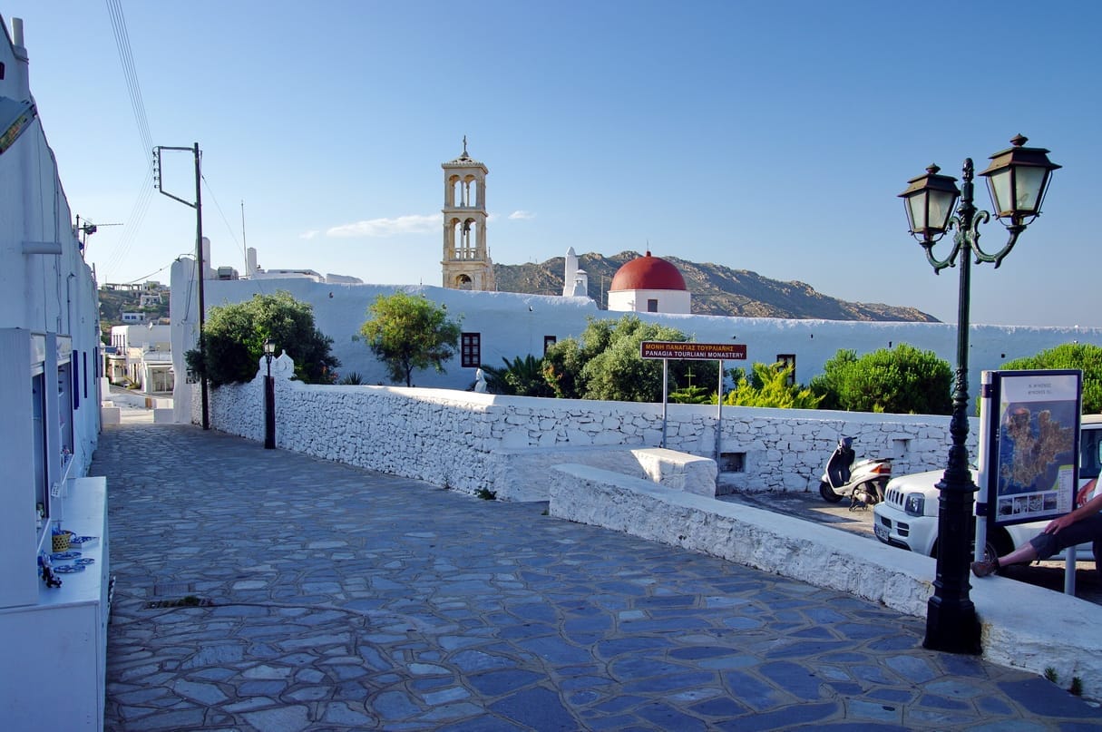 Calle empedrada en Ano Mera con edificios tradicionales encalados, árboles verdes y el campanario del Monasterio de Panagia Tourliani bajo un cielo azul despejado.