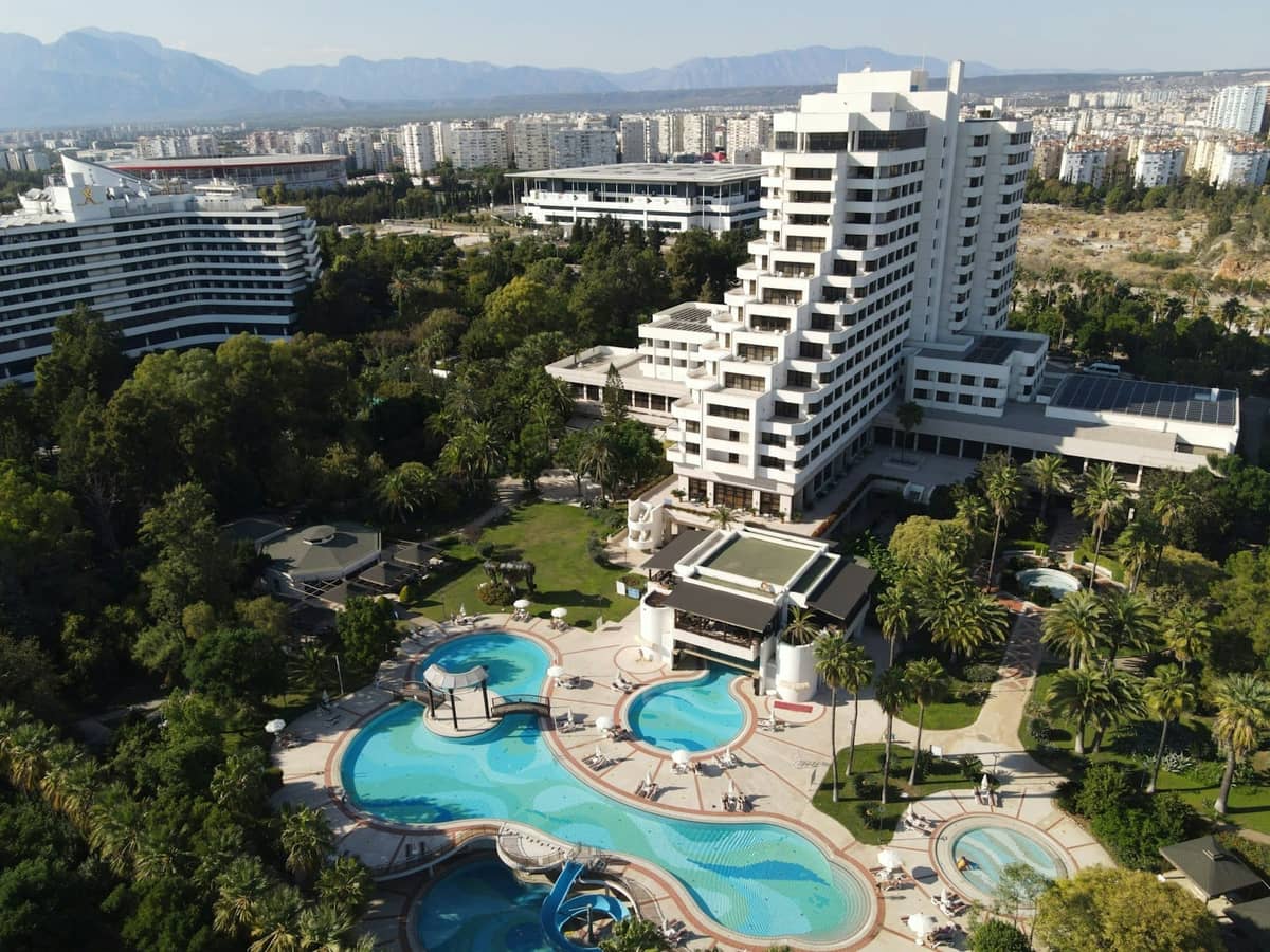 Aerial view of a luxury resort in Antalya with multiple pools, sun loungers, and modern hotel buildings surrounded by greenery.