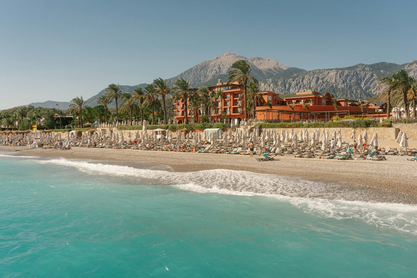 Um resort à beira-mar em Antalya com fileiras de espreguiçadeiras, palmeiras, um grande edifício e montanhas ao fundo sob céu limpo.
