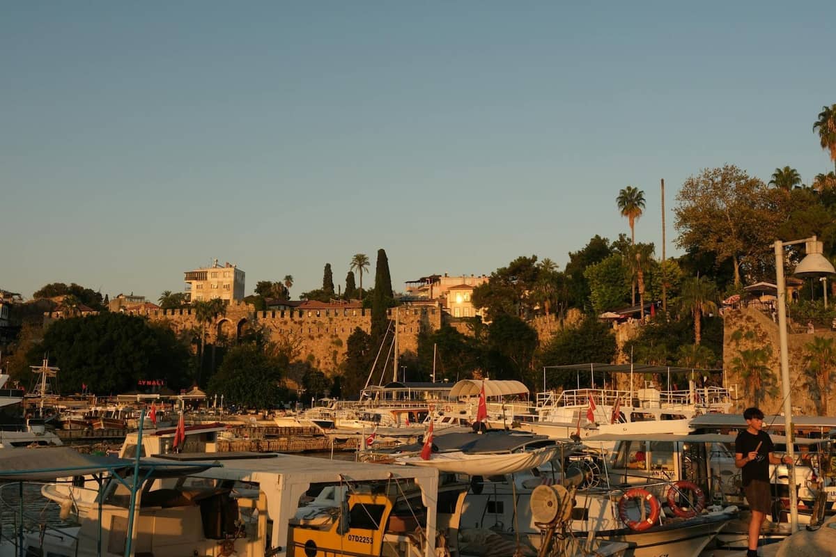 Antalya harbor at sunset with boats docked and historic city walls and greenery in the background, capturing the area’s coastal charm.