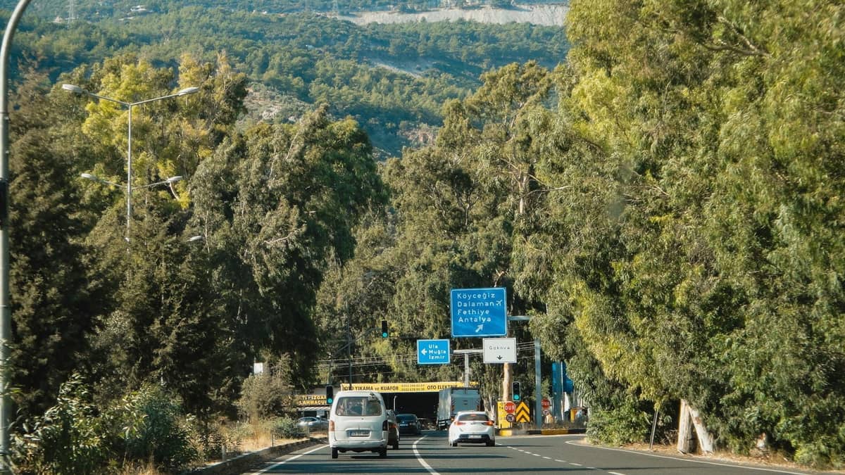 Cars driving on a highway near Antalya with a road sign showing directions to city destinations surrounded by lush trees and hills.