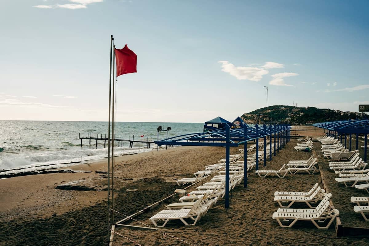 Rows of empty sun loungers and blue sunshades on a long sandy beach with gentle waves and a red flag, east of Antalya.
