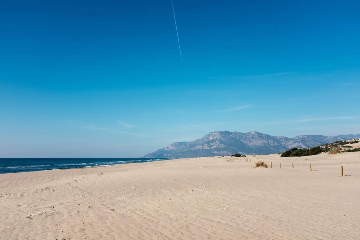 Wide sandy beach with distant mountains, blue sky, and gentle waves; natural, open, and inviting landscape.