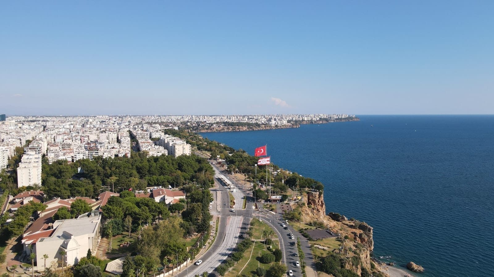 Vista aérea de Antalya com a orla, o silhueta da cidade, avenidas principais, áreas verdes e uma grande bandeira turca sob um céu azul límpido.