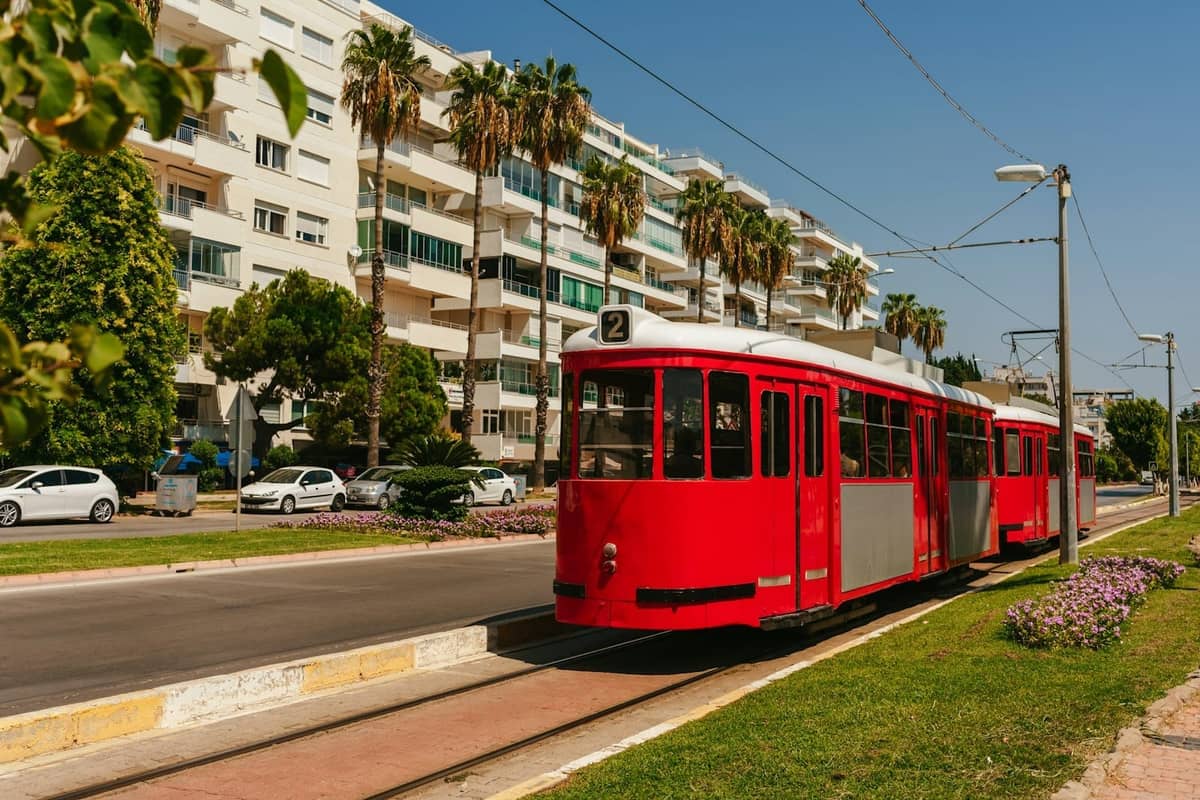 A bright red tram traveling along tracks in a sunny Antalya street lined with palm trees and modern apartment buildings.