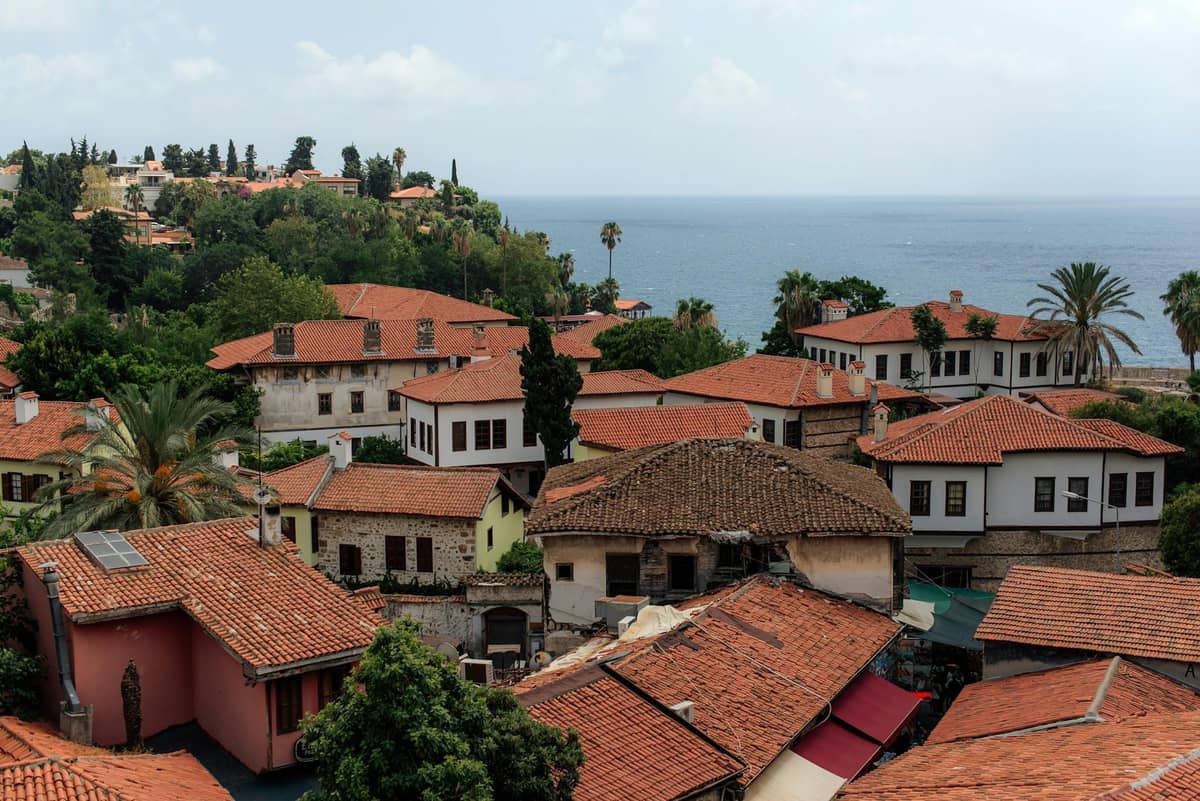 A panoramic view of Antalya's historic neighborhood with red-tiled roofs, greenery, and the sea in the background.