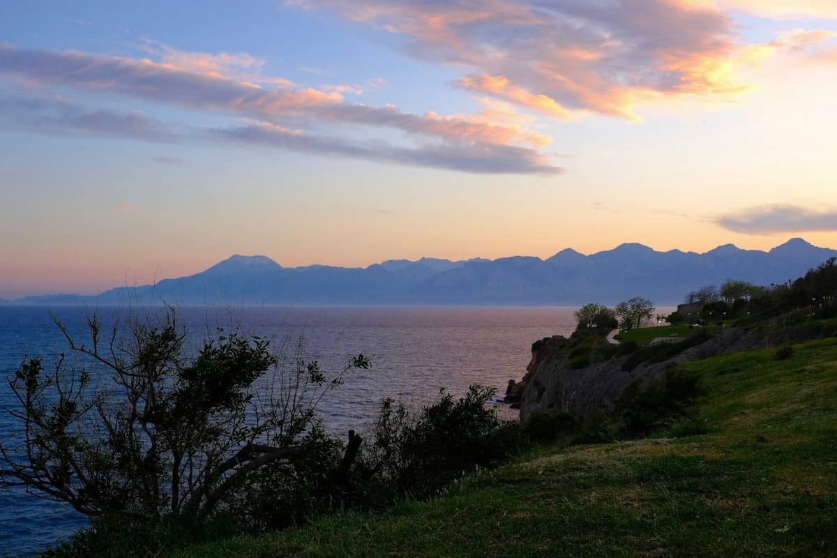 Cliffside park in Antalya at sunset with sea views, silhouettes of trees, and distant mountains under a colorful sky.