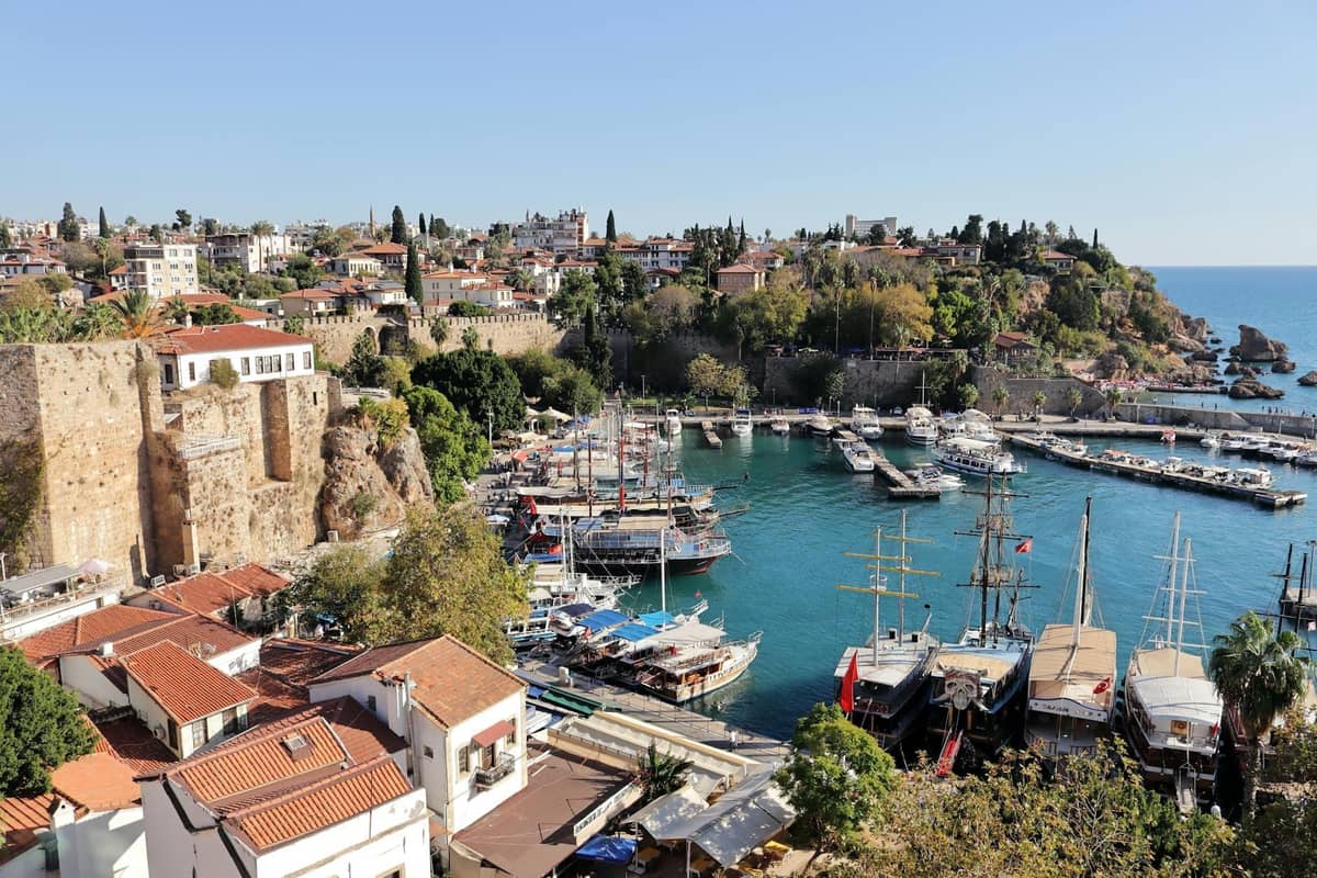 Antalya's old harbor on a bright sunny day, traditional wooden boats in turquoise water, historic stone walls, and red-roofed houses under a clear summer sky.