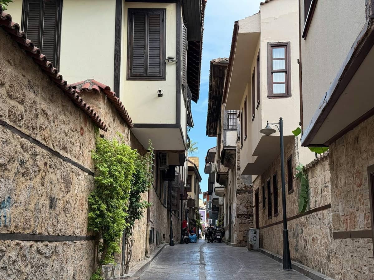 Narrow stone-paved street lined with historic Ottoman-style houses in Kaleiçi, Antalya, with plants and bicycles in the distance.