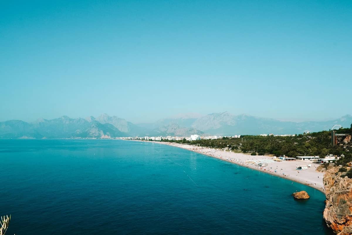 Wide view of Konyaaltı Beach in Antalya with turquoise sea, sandy shoreline, distant mountains and scattered sunbathers, under a clear blue sky.