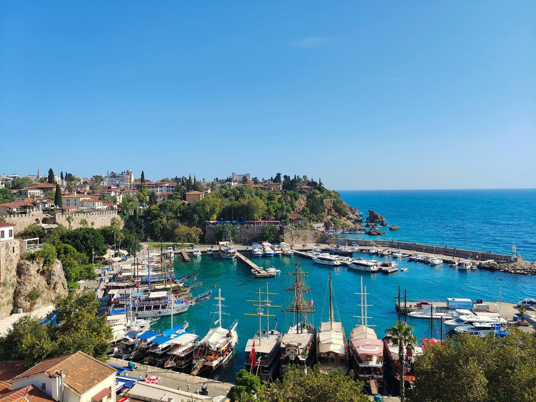 Vista panoramica della Marina di Antalya con acque turchesi, edifici storici della città vecchia, vegetazione lussureggiante e barche da tour ormeggiate lungo il porto semicircolare sotto un cielo azzurro.