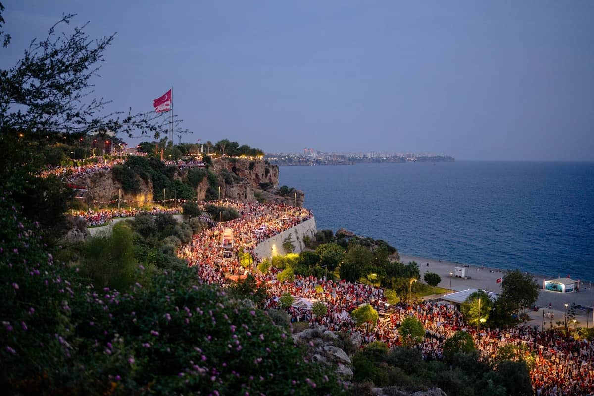Crowds gathering along Antalya's dramatic seaside cliffs at dusk, with bright lights and a festive summer atmosphere by the water.