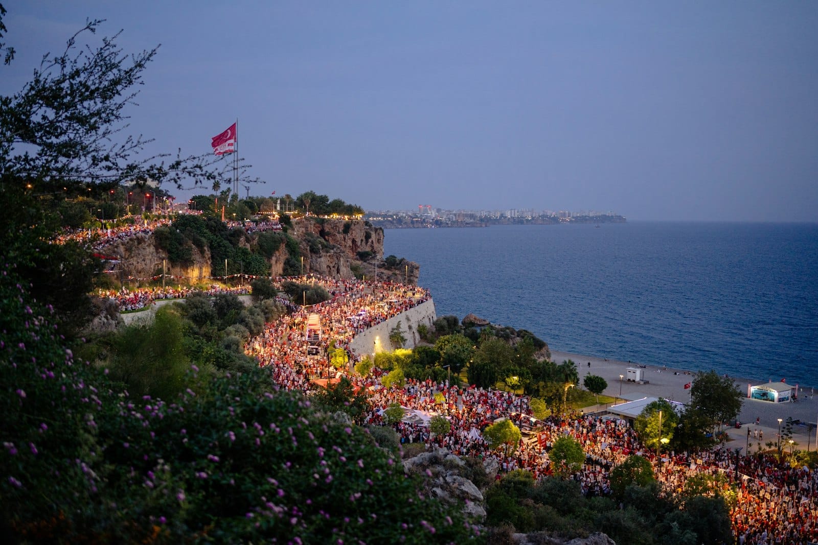Grande multidão reunida nos penhascos de Antalya ao entardecer com vista para o mar, luzes da cidade começando a brilhar, atmosfera animada de fim de tarde com a bandeira turca visível.