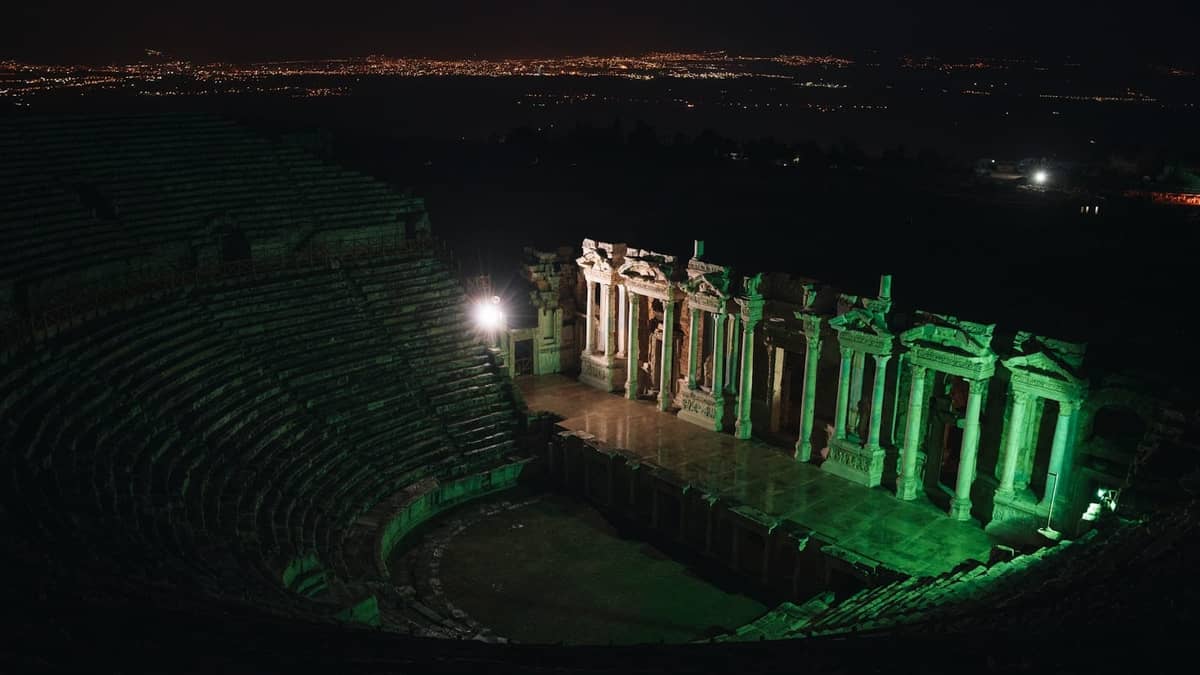 Night view of a grand Roman amphitheater brightly lit, with city lights in the distance, suggesting an evening cultural event.
