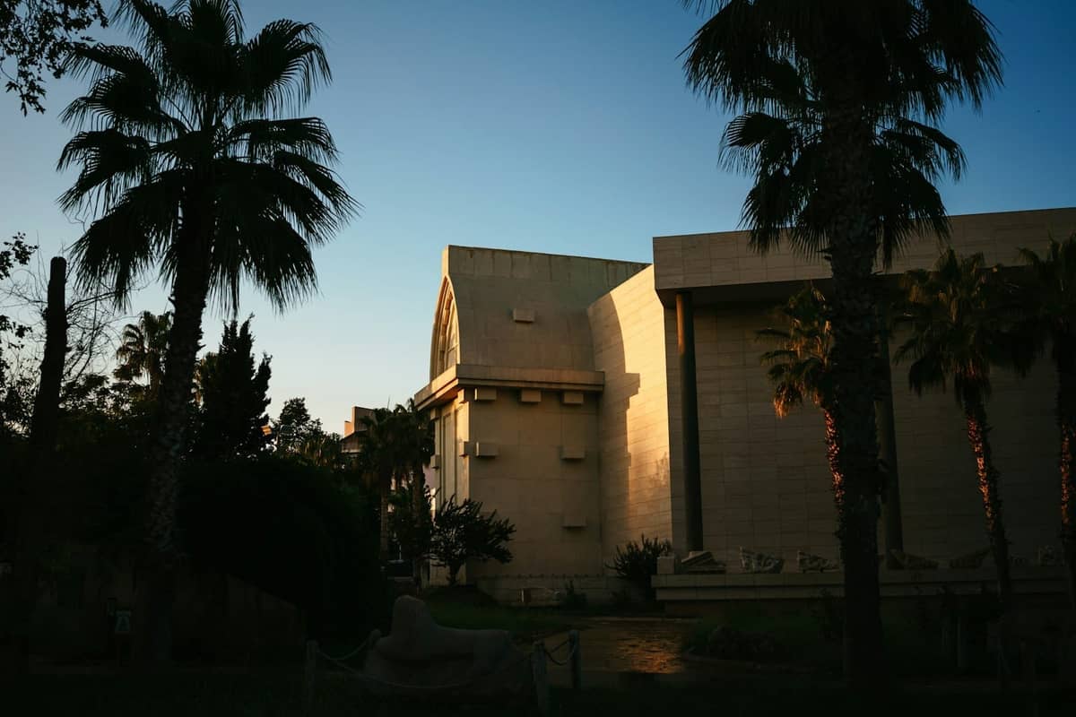 Exterior view of Antalya Museum building partially in shadow, surrounded by palm trees in late afternoon light.