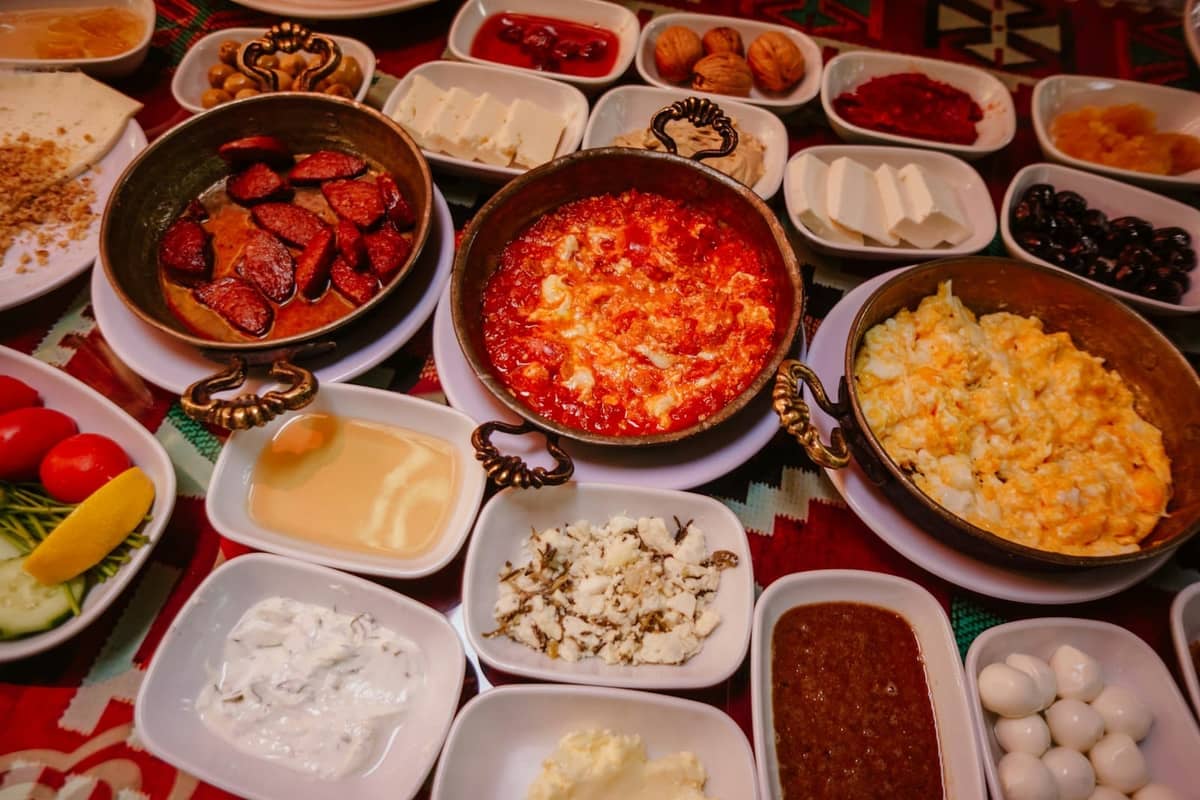 A colorful Turkish breakfast spread with small dishes of eggs, cheese, olives, breads, and spreads arranged on a patterned tablecloth.