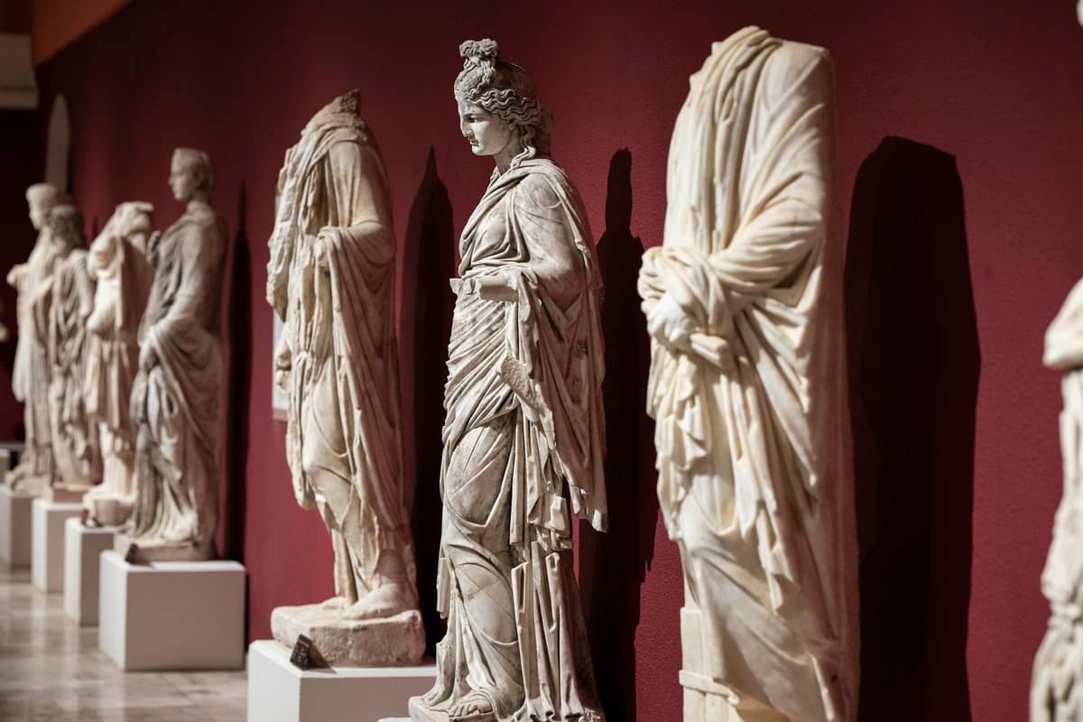 A row of ancient marble statues displayed inside a museum gallery against a red wall, well-lit and detailed.