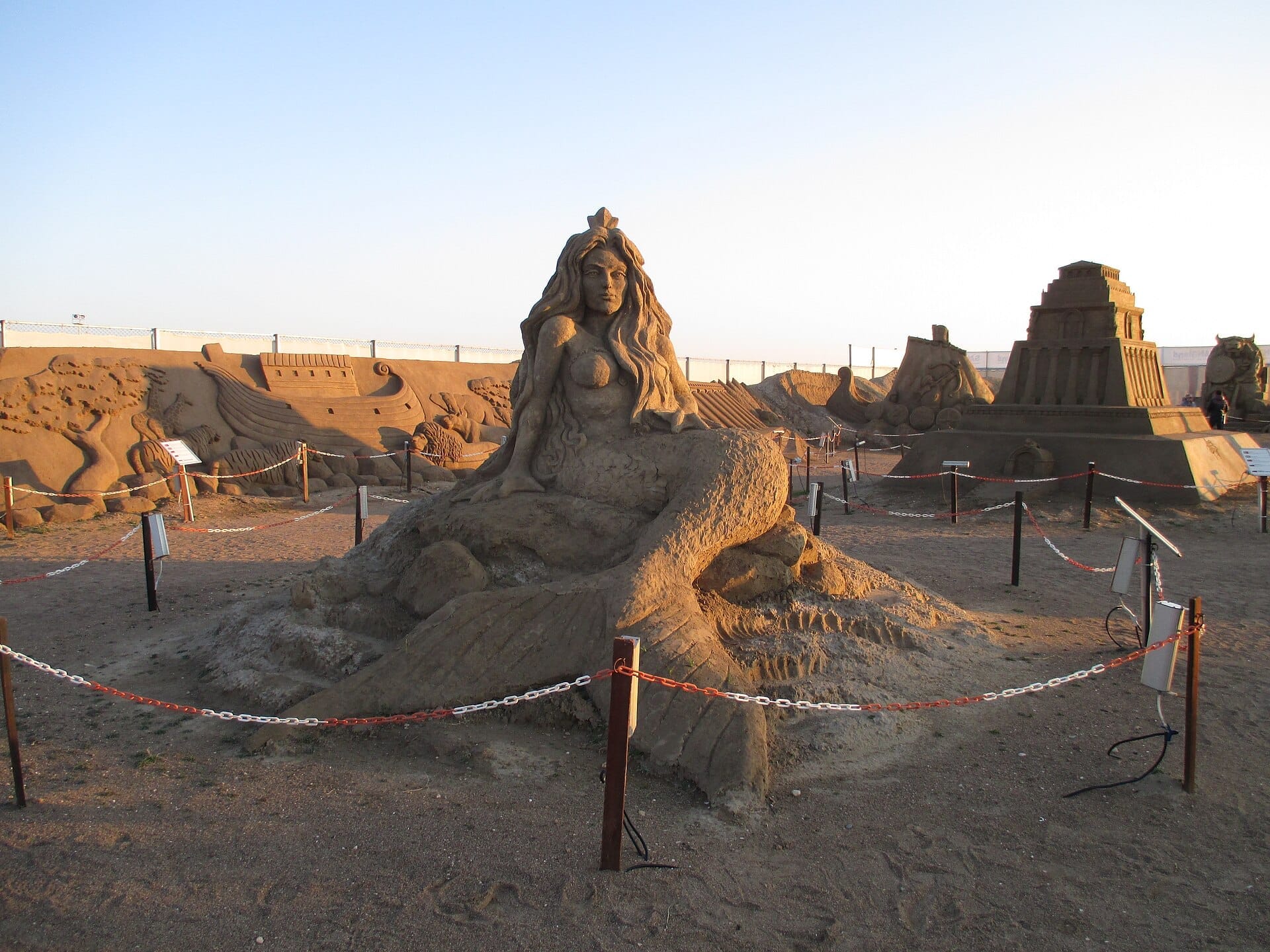 Grandi sculture di sabbia, tra cui una sirena e strutture antiche, esposte al Sandland Antalya sotto un cielo sereno serale.
