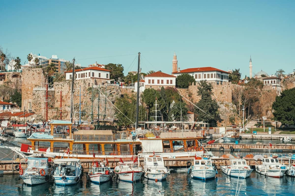 View of Antalya marina with boats docked in front of the old town and historic stone buildings under a clear blue sky.