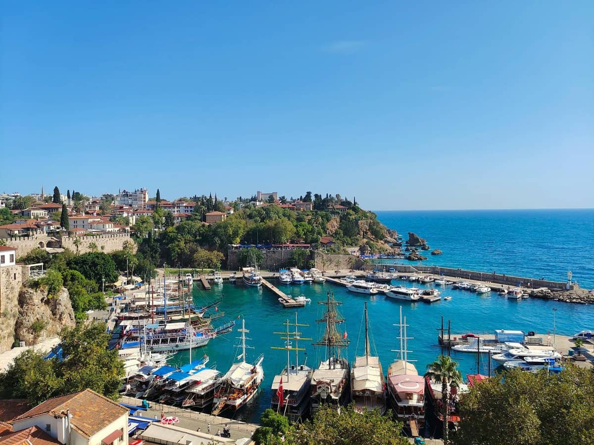 Bright daytime view of Antalya's Old Harbor with moored boats, historic buildings on the cliffs, and clear blue sea, bustling with activity.