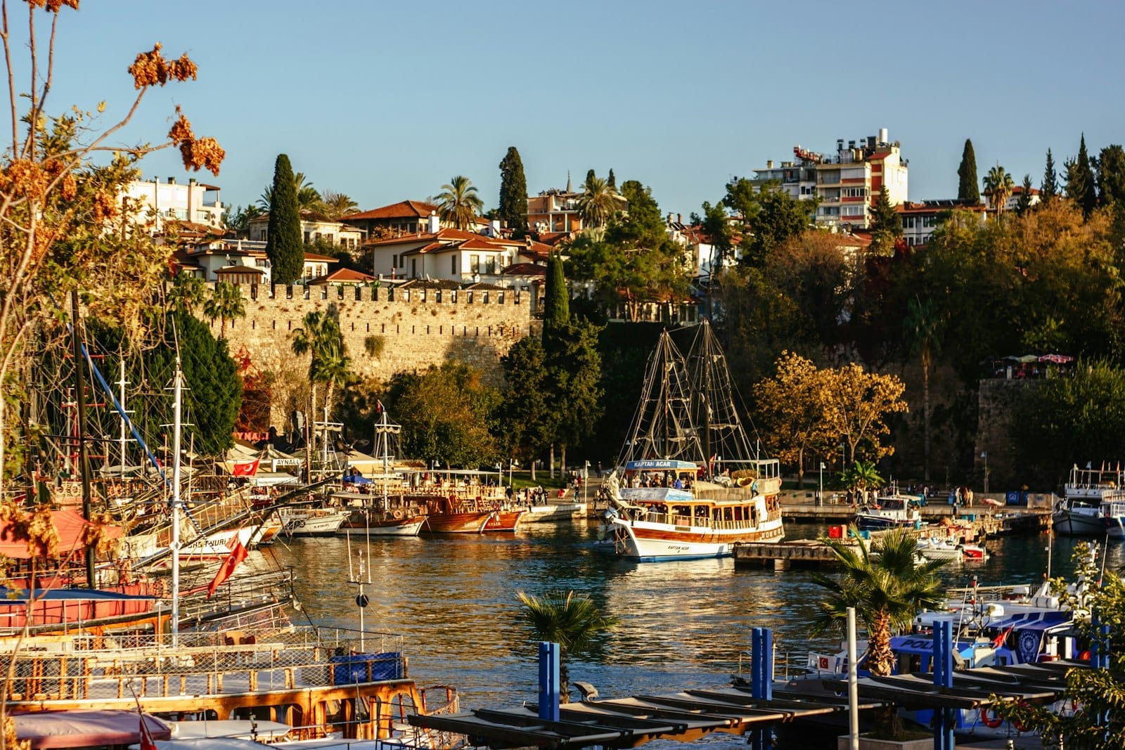 Vista panorâmica do porto da Cidade Velha de Antalya com barcos coloridos, antigas muralhas e árvores frondosas sob um céu limpo, com todo o charme típico do Mediterrâneo.