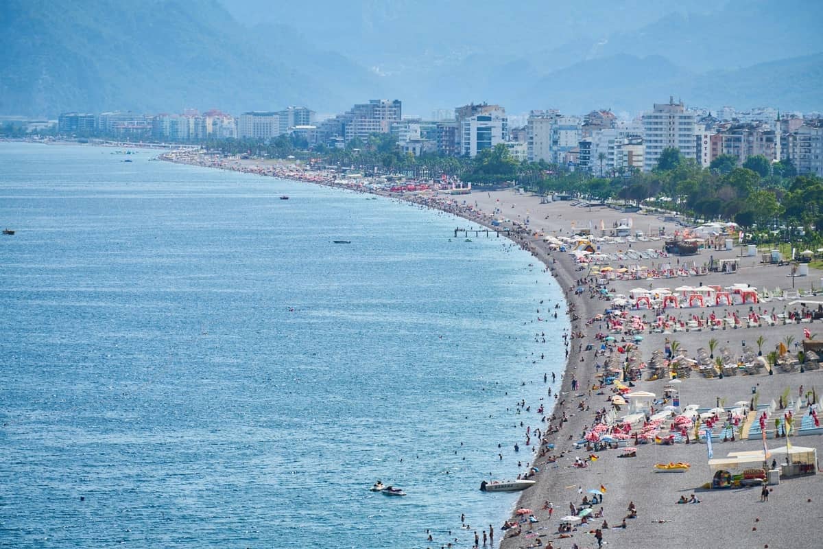 Wide view of Antalya's city beach packed with people, sun umbrellas, and high-rise buildings during summer peak season.