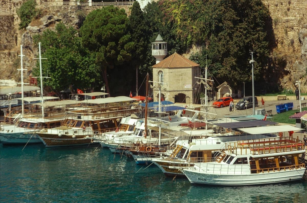 Several tour boats docked along Antalya's old harbor, with stone buildings, trees, and cliffs in the background under bright sunlight.
