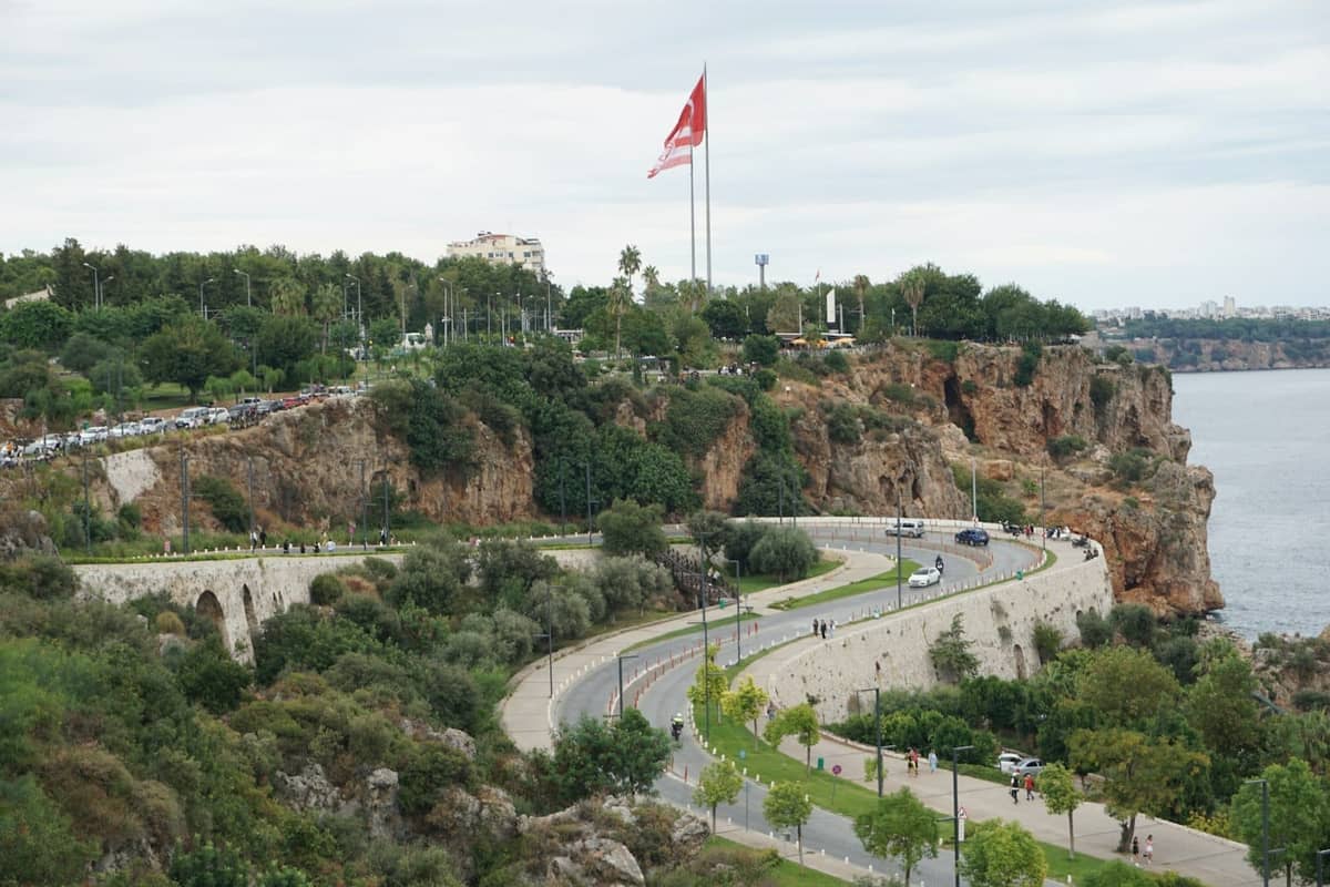 Clifftop park overlooking the sea with pathways, greenery, people walking, and a large Turkish flag near Antalya’s coastline.