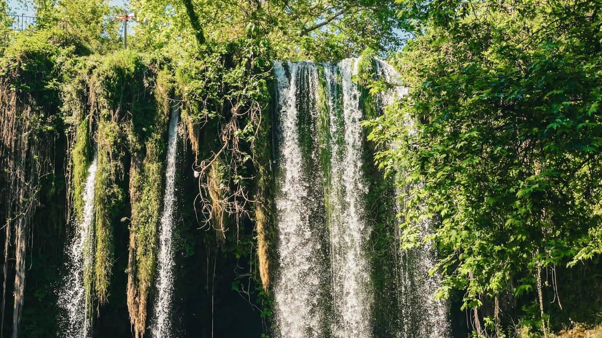 A tall, scenic waterfall surrounded by lush green vegetation in bright daylight, with water cascading into a shaded pool below.