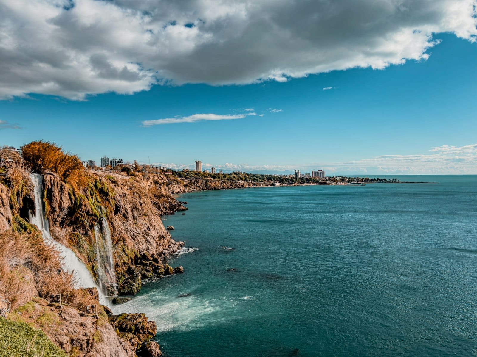 Vista ampla das Cachoeiras Düden Inferior de Antalya despencando no azul do Mar Mediterrâneo, com o silhueta da cidade e nuvens dramáticas ao fundo.