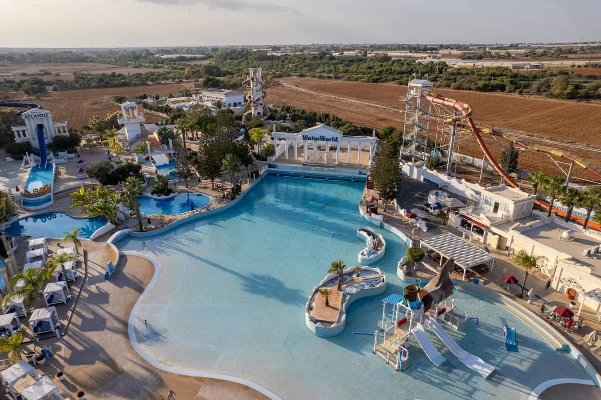 Aerial view of a large water park with pools, water slides, and play areas, surrounded by loungers and palm trees, under a partly cloudy sky.