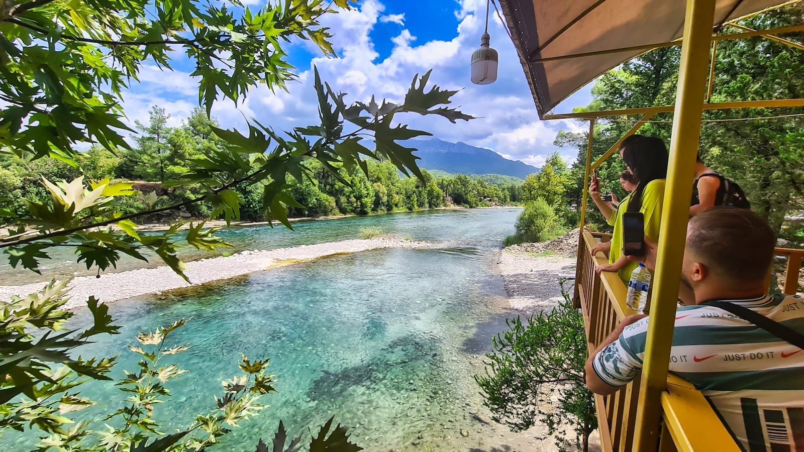 Crianças e adultos aproveitam um terraço à beira do rio com água cristalina, árvores verdes e montanhas ao fundo sob um céu azul intenso perto de Antalya.