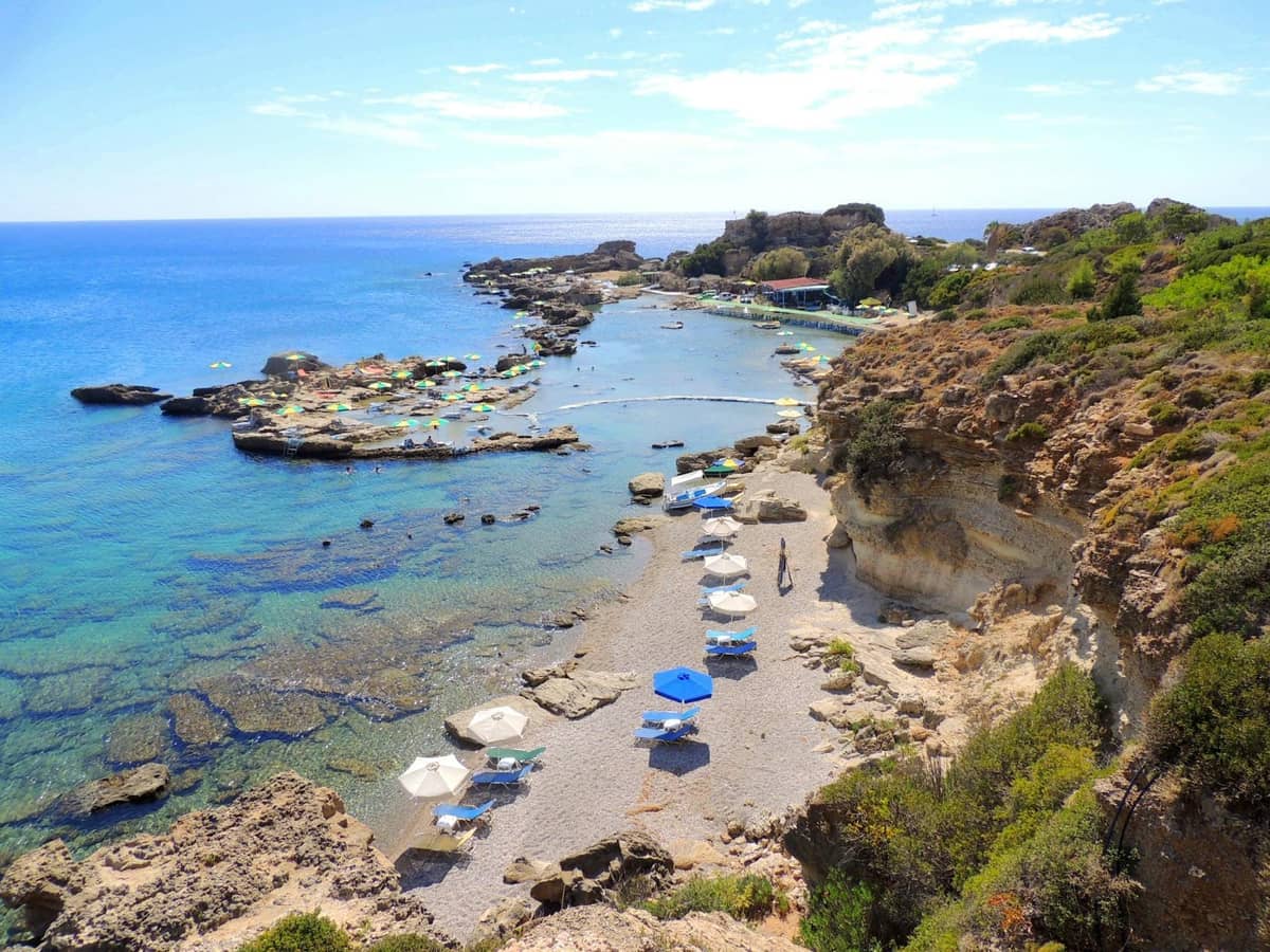 Rocky cove with clear turquoise water, scattered umbrellas, sunbeds, and surrounding greenery at Anthony Quinn Bay in Rhodes.