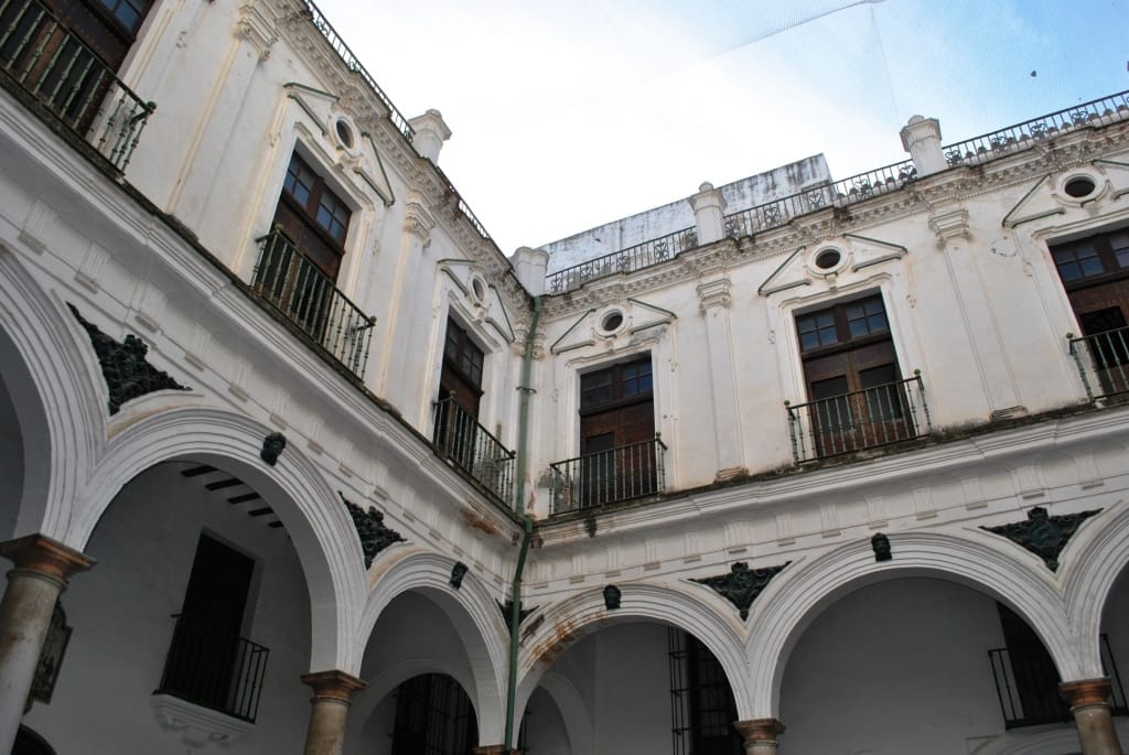 Il cortile interno dell'Antiguo Hospital de Mujeres a Cádiz, con archi barocchi, colonne e facciate bianche consumate dal tempo sotto un cielo azzurro.