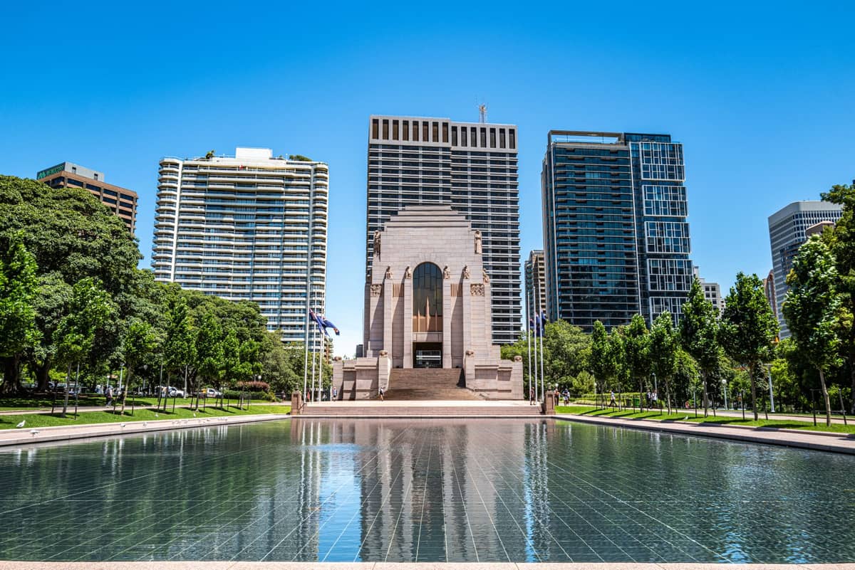 Vista frontal del Memorial ANZAC en Sídney, fotografiado con su reflejo en el Estanque de Reflexión, rodeado de árboles y edificios modernos en un día soleado.
