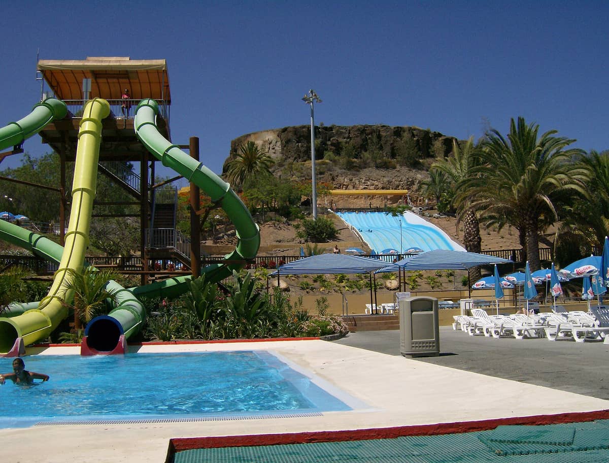 Вид на аквапарк Aqualand Maspalomas на Гран-Канарии: высокие горки, бассейн, пальмы и шезлонги под ясным голубым небом.