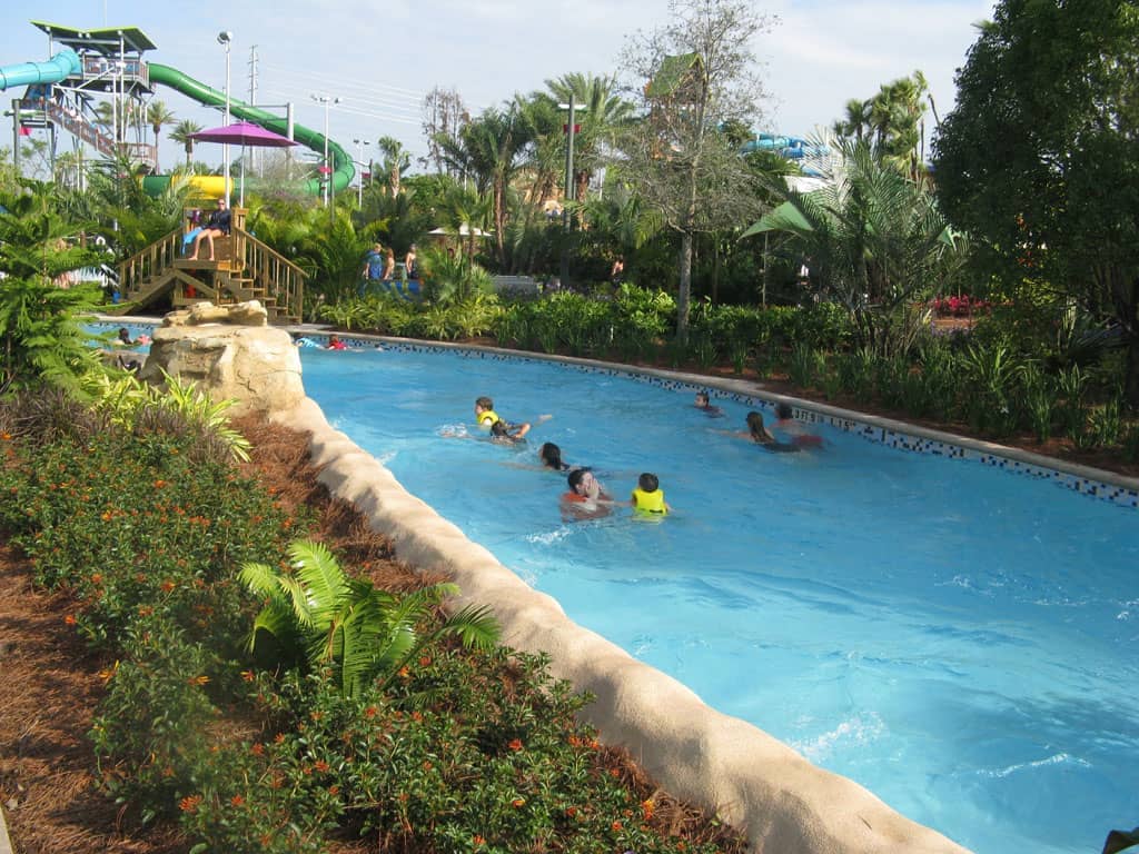 Familias flotando en el río lento panorámico de Aquatica Orlando, rodeadas de exuberante vegetación y vibrantes toboganes de agua bajo un cielo parcialmente nublado.