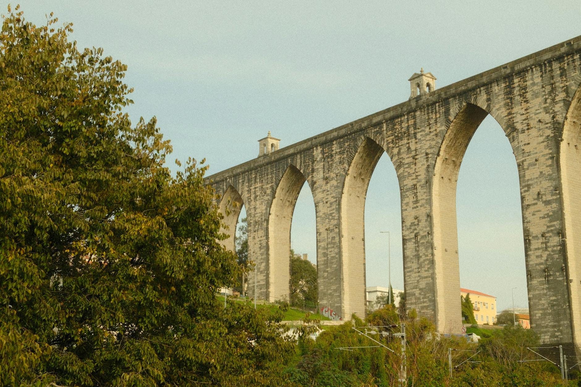 Vista dell'Acquedotto delle Águas Livres a Lisbona, con le imponenti arcate gotiche in pietra che si stagliano sopra le chiome degli alberi su uno sfondo di cielo azzurro.