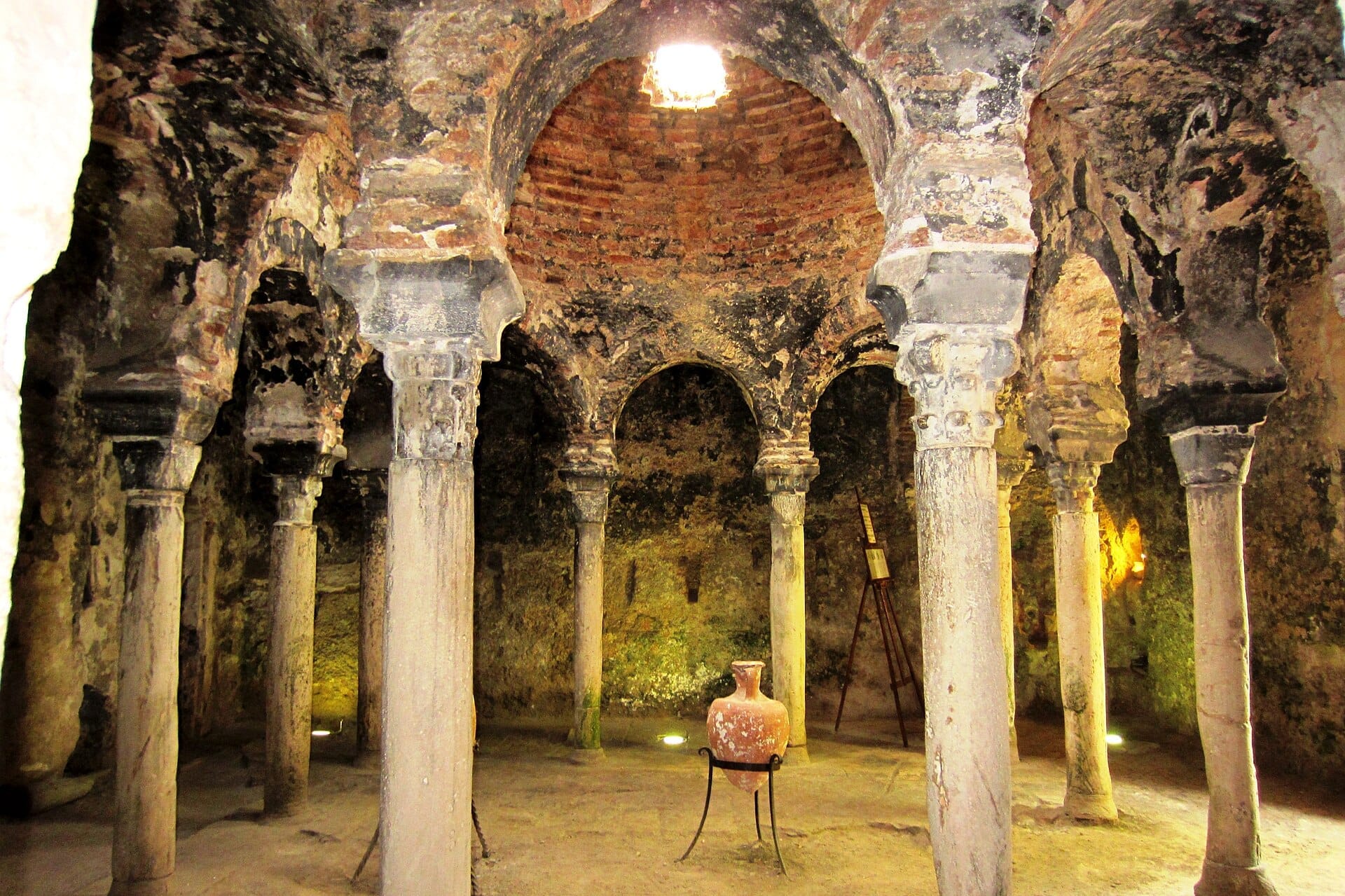 Interno dei Bagni Arabi di Palma, con antiche colonne in pietra che sorreggono archi in mattoni e un'urna centrale in argilla sotto luce naturale.