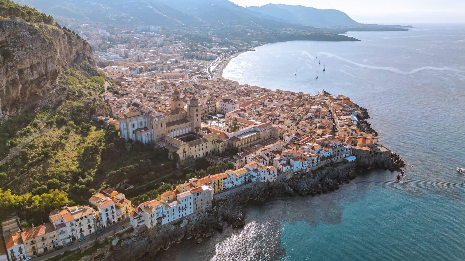 Luftaufnahme von Cefalù mit seiner Kathedrale, dem historischen Zentrum und dem dramatischen Felsenhintergrund neben dem blauen Tyrrhenischen Meer.