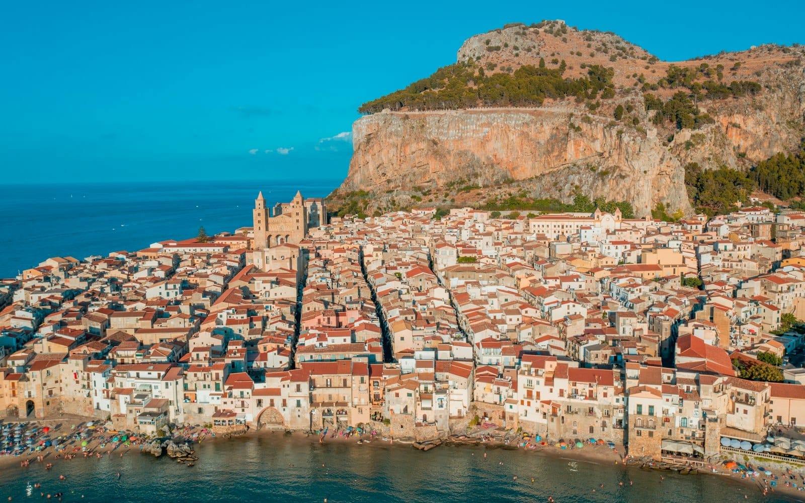 Luftaufnahme von Cefalù mit seiner mittelalterlichen Kathedrale, dem kompakten historischen Zentrum, dem umliegenden Meer und dem dramatischen Felsberg im Hintergrund unter strahlend blauem Himmel.